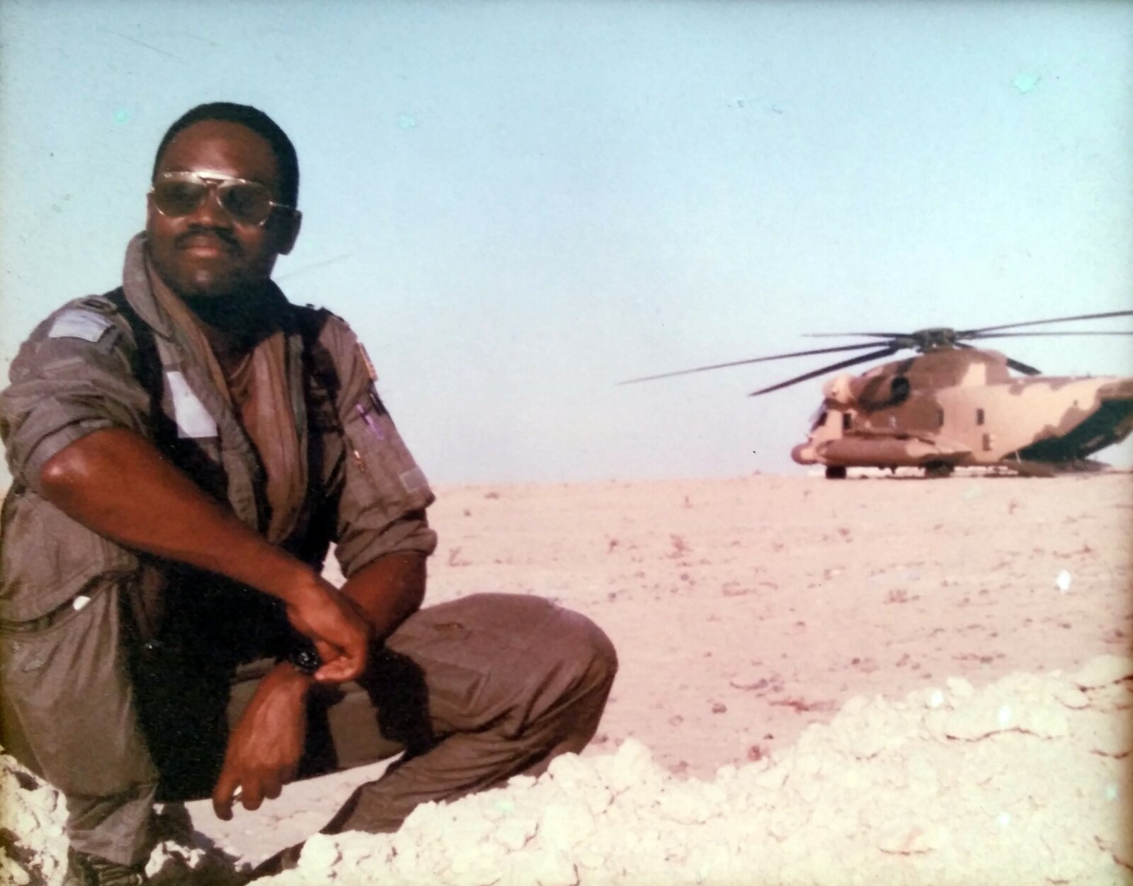 Donald Mitchell in a flight suit, crouching in the desert with a desert-camouflaged MH-53J Pave Low helicopter behind him, Desert Storm, 1991.